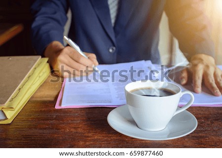 Black coffee on a wood table in the morning and a file with a male businessman signing a document at work.