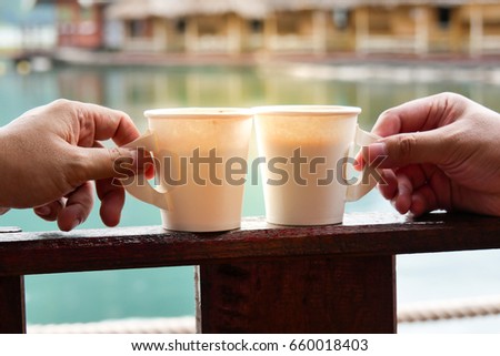 two hands holding cups of coffee, chocolate, tea  with lake and hut background