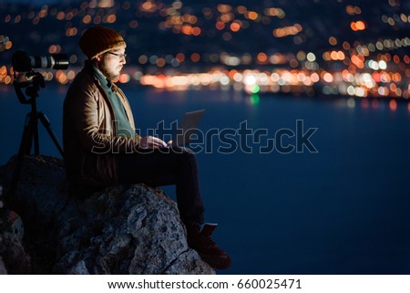 Young attractive photographer sitting on a rock and working on laptop with perfect view of sea, port, night city and sky full of clouds on background. Camera on tripod beside him.