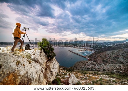 Young attractive photographer standing on a rock and making panoramic photos of night city with camera on tripod at sunset. Sea, port and beautiful sky full of clouds on background.