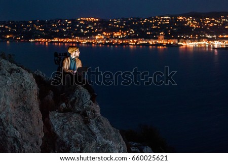 Young dreamy photographer sitting on a precipice, working on laptop and looking at a perfect view of sea, port, night city and sky full of clouds on background. Camera on tripod beside him.