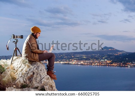 Young attractive photographer sitting on a rock and working on laptop with perfect view of sea, port, night city and sky full of clouds. Camera on tripod beside him.