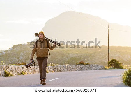 Young attractive photographer wearing casual clothes, yellow cap and glasses walking on desert road with tripod on his shoulder and camera in hand. Mountains on background