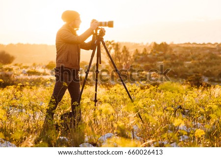 Portrait of young photographer making photos of nature with camera on tripod at sunset. Sunny day.