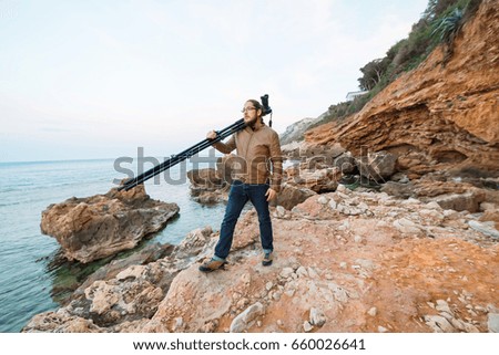 Young attractive male photographer with beard and long hair wearing casual clothes is standing on the rock with his camera on tripod and looking into the sea.