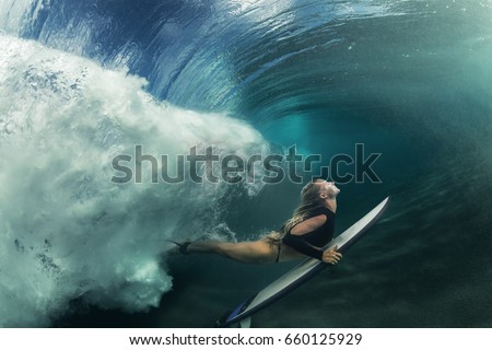 A blonde surfer girl underwater doing duck dive holding surfing board left behind air bubbles in blue water background under big ocean wave