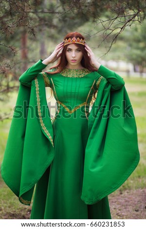 A beautiful, young, red-haired girl in a green medieval dress with long sleeves, with a golden crown, standing on the grass, in a fairy forest. Fantastic photosession. A model with clean skin.