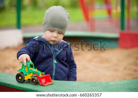 Portrait of cute toddler boy sitting on the ground and playing with toy tractor and sand in the park. Child walking outdoors. Lifestyle.