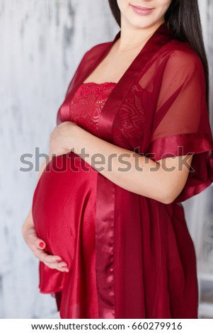 Close up photo of young pregnant happy woman in trendy red peignoir smiling and touching her belly with hands on white background.