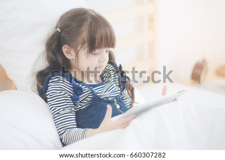 Cute asian little girl enjoy watching cartoon on smart tablet while sitting on bed in kid bedroom at home.