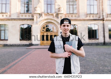young man stands at the entrance to his university and holds a notebook in his hands. A smiling student on the background of his college campus. Student life. Look at the camera