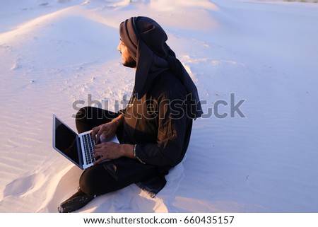 Joyous male Arab sits on sand at computer. young man engaged in scientific work or writes article typing on keyboard of gadget, relaxes sitting in silence of bottomless desert against blue sky on