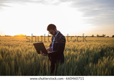 Young man in black suit using laptop in wheat field, during beautiful sunset
