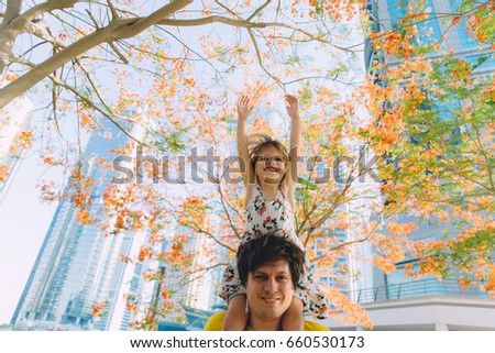 Little girl sitting on her father's shoulders in the city with trees in blossom and skyscrapers on the background