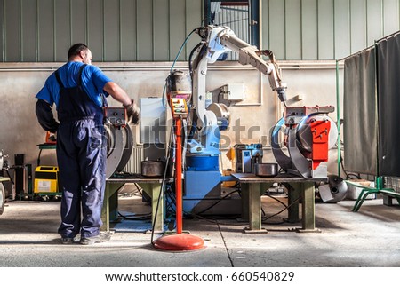 Man and robotic machine work together inside industrial building. The mechanical arm performs welds on metal components assisted by a worker.