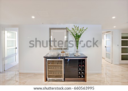 Glasses on the wine drawer cabinet in the hallway of a modern house interior, the cabinet has bottle store like a fridge. there is a green plant beside the wall and ceiling