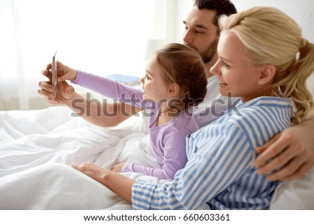 people, family and technology concept - happy mother, father and little girl taking selfie with smartphone in bed at home