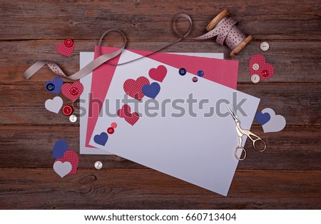 Selection of scrapbooking papers, card, shapes like hearts, buttons, ribbon on a bobbin and scissors on a wooden background with a vintage feel. With space for text. 