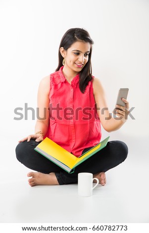 Pretty Indian/Asian College Girl studying from book and using Smartphone while sitting over white background
