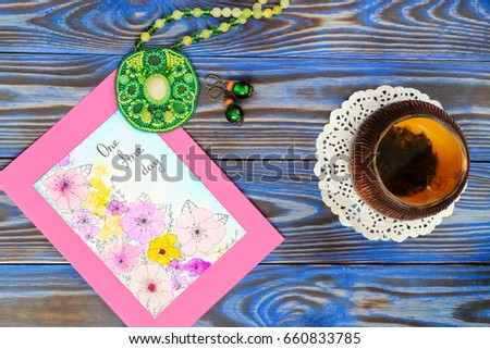 Ceramic bowl piala cup with green tea with jasmine and picture card with inscription "One fine day", green pendant made from a seed bead and earrings on blue boards background. Happiness Still life