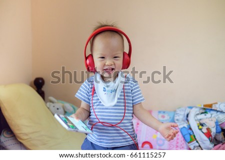 Cute little Asian 18 months / 1 year old baby boy child listening music with headphones from smartphone, Kid stand & Play on bed in bedroom at home, leisure & technology & internet addiction concept