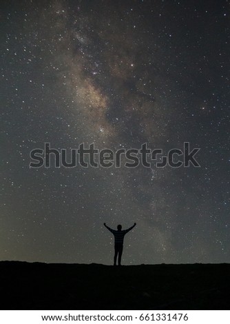 Night colorful landscape with Milky Way, yellow city lights and silhouette of a standing sporty man with raised up arms on the mountain. Universe. Travel background with purple sky full of stars  