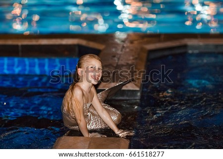 Blonde girl in a fantasy mermaid costume in a swimming pool. Cute little girl sitting poolside in a mermaid suit. A child of five or six years with a mermaid's tail bathes, smiles. Night photo.