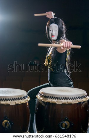 drummer girl in white demon mask with drumsticks, studio concert shot on a dark background.