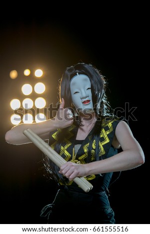 drummer girl in white demon mask with drumsticks showing ok sight, studio concert shot on a dark background and soffit lights.