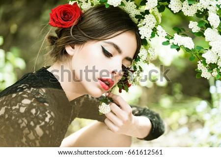 girl with fashionable makeup and red lips, has rose flower in hair hispanic or spanish style in black dress at white spring or summer blossom on natural background