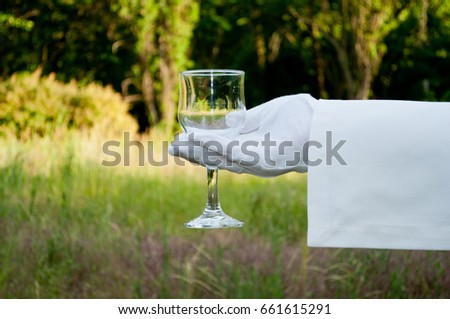 Hand of the waiter in a white glove and with a white napkin holding a glass glass with water for wine on a blurred background of nature green bushes and trees