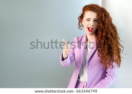 Portrait of successful happy beautiful business woman with red - brown hair and makeup in pink suit with thumbs up and wink. looking at camera with toothy smile,  studio shot on gray background.