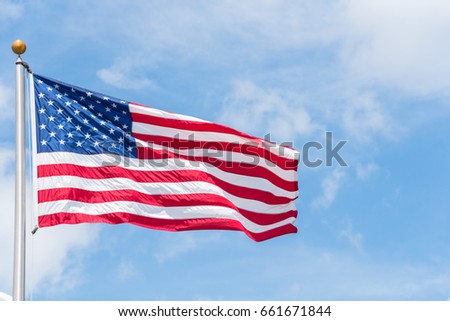 Large American flag waving on flag pole with cloud blue sky. Windy and sunny day with waving star and striped flag blowing/flowing. Ruffled USA flag. Room for text.