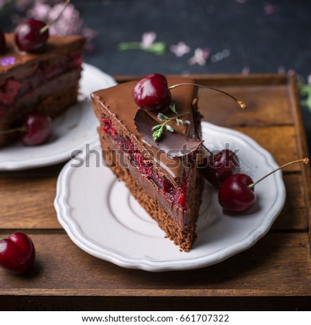 A piece of chocolate cherry mousse cake is decorated with lilac flowers and fresh cherries on a wooden box on a wooden dark background.  Dark food photo
