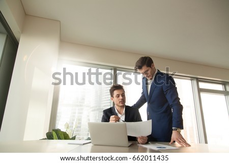 Serious boss talking to employee at workplace, discussing document, reporting on project, showing presentation, demonstrating task results, team leader checking work of subordinate manager