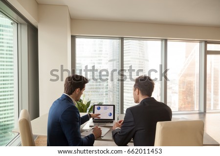 Two men discussing company growth, looking at rising graph on laptop screen, using easy accounting software for small business, stock traders analyzing market, pointing at charts on monitor. Rear view Royalty-Free Stock Photo #662011453