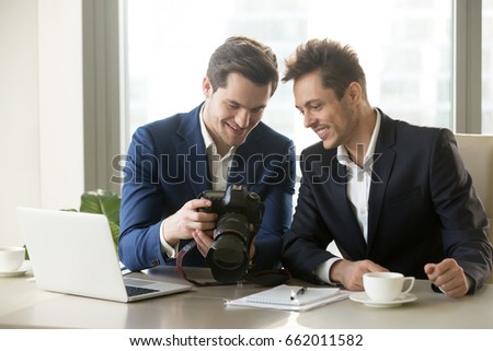 Happy businessman holding new professional camera, showing photographs to smiling colleague, partners viewing discussing pictures of business object, satisfied client looking at photos for project ads