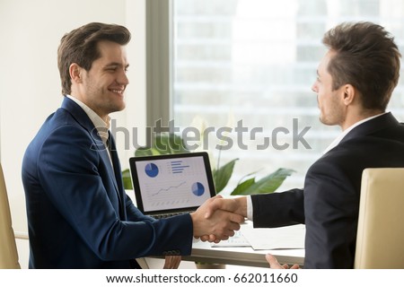 Two smiling businessmen handshaking, sitting at desk with charts graphs on laptop screen, partners shaking hands after successful financial negotiations, satisfied with result, growth, help, support Royalty-Free Stock Photo #662011660