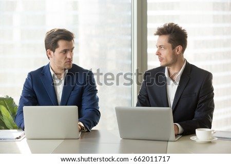 Two confident businessmen sitting at office desk with laptops, looking at each other with hate dislike, rivals accepting challenge, business competition, team rivalry at work, competitors behavior Royalty-Free Stock Photo #662011717