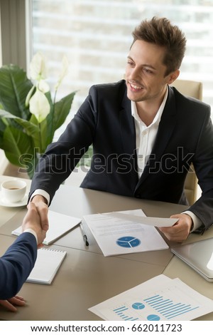 Vertical portrait of happy businessman handshaking at meeting over office desk with graphs charts, holding financial document, offering contract, manager and client shaking hands, making good deal
