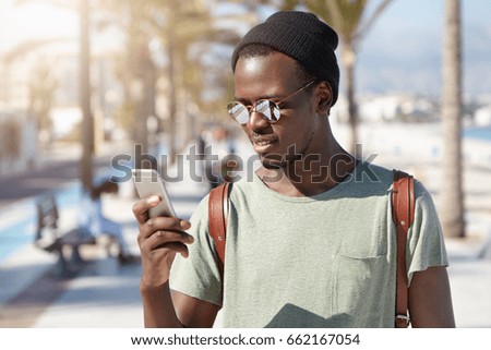 Headshot of African male wearing stylish hat and sunglasses holding smartphone typing messages using free internet connection having holidays abroad isolated over big city background. Technologies