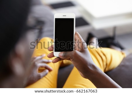 Back view of hipster guy lying at cozy sofa holding modern smartphone with blank screen for your text messaging and looking photos online. Cropped view of dark-skinned male holding mobile phone