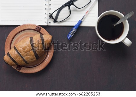 croissant with coffee cup ,eye glass,pen,notebook on dark wooden table,coffee time