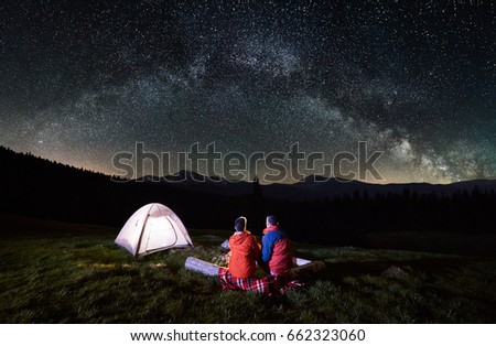 Night camping in the mountains. Rear view of romantic couple tourists have a rest at a campfire near illuminated tent under amazing night sky full of stars and milky way. Low light