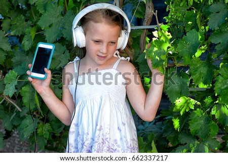 Happy beautiful little girl with headphones listening  to music  in her cell phone  in the summer park. Blank screen on phone . Summer holidays. Children and technology.  