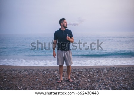 Vape. A brutal young man with a beard smoking an electronic cigarette by the sea at sunset. Lifestyle.