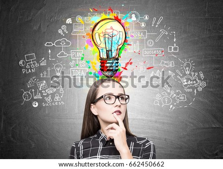 Portrait of a nerdy girl in glasses standing near a blackboard with a business scheme and a colorful light bulb above her head