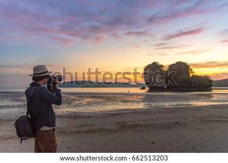 Young Asian man photographer taking photo of summer landscape with beautiful sunset sky at Ao Nang Beach, famous tourist attraction and travel destination of Krabi Province, Thailand
