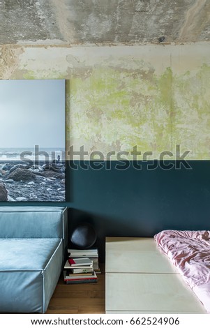 Wooden bed with rose linens on the background of the shabby wall. On the left there is a sofa with a big photo over it, piles of books and a black speaker. Closeup. Vertical.