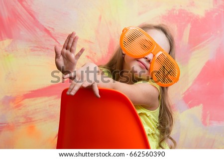 child. happy child or small girl sitting on orange chair on colorful abstract background in summer glasses and tshirt with long blond hair